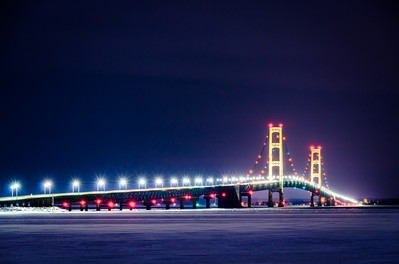 Mackinac Bridge is seen at night, lit up & reflecting in the dark water below as it stretches over it. The Mackinac Bridge is a suspension bridge spanning the Straits of Mackinac, connecting the Upper and Lower peninsulas of the U.S. state of Michigan.